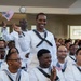 A group of U.S. Navy sailors in white dress uniforms are gathered in what appears to be an indoor ceremony hall. A central figure is standing and smiling broadly while waving a small American flag, with another sailor behind also waving a flag. The atmosphere appears celebratory, consistent with a naturalization or graduation ceremony.