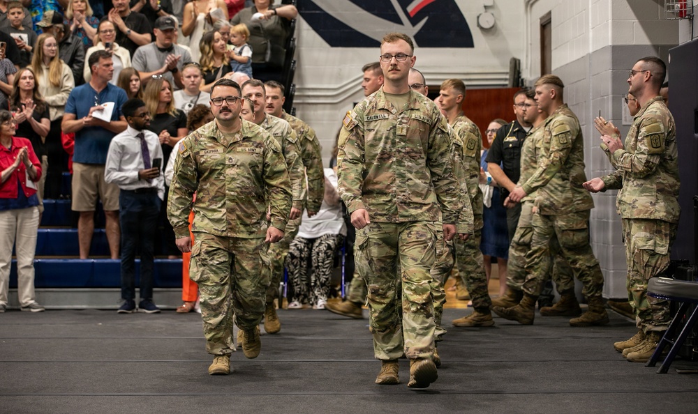 A group of U.S. Army soldiers in OCP (Operational Camouflage Pattern) uniforms march in formation through what appears to be a gymnasium or arena filled with civilian spectators. The crowd is applauding, suggesting this is a formal military ceremony such as a deployment sendoff, homecoming, or commissioning event. Two soldiers lead the formation prominently in the foreground.