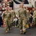 A group of U.S. Army soldiers in OCP (Operational Camouflage Pattern) uniforms march in formation through what appears to be a gymnasium or arena filled with civilian spectators. The crowd is applauding, suggesting this is a formal military ceremony such as a deployment sendoff, homecoming, or commissioning event. Two soldiers lead the formation prominently in the foreground.