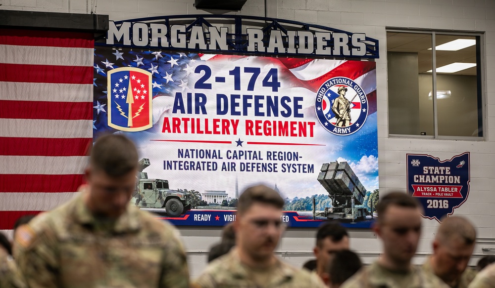 The image shows a military ceremony or gathering for the 2-174 Air Defense Artillery Regiment of the Ohio Army National Guard, held inside what appears to be a school gymnasium (Morgan Raiders). Multiple soldiers in camouflage uniforms are assembled in the foreground, with a large promotional banner visible in the background. The event is related to the unit's mission supporting the National Capital Region Integrated Air Defense System.