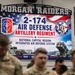The image shows a military ceremony or gathering for the 2-174 Air Defense Artillery Regiment of the Ohio Army National Guard, held inside what appears to be a school gymnasium (Morgan Raiders). Multiple soldiers in camouflage uniforms are assembled in the foreground, with a large promotional banner visible in the background. The event is related to the unit's mission supporting the National Capital Region Integrated Air Defense System.
