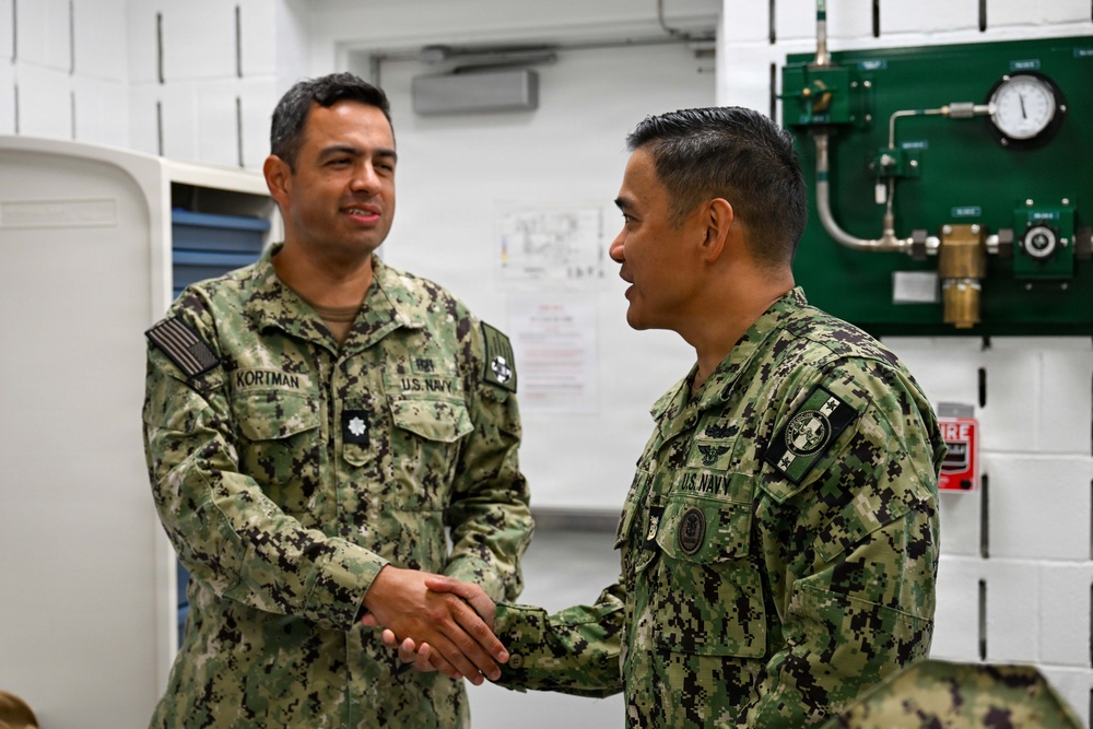 Two U.S. Navy personnel in AOR-2 (woodland digital camouflage) uniforms are shaking hands in what appears to be a military facility or training room. The individual on the left is identified by name tape as 'KORTMAN' and wears a Lieutenant Commander oak leaf cluster rank insignia. Both wear U.S. Navy branch tapes and various qualification badges on their uniforms.