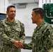 Two U.S. Navy personnel in AOR-2 (woodland digital camouflage) uniforms are shaking hands in what appears to be a military facility or training room. The individual on the left is identified by name tape as 'KORTMAN' and wears a Lieutenant Commander oak leaf cluster rank insignia. Both wear U.S. Navy branch tapes and various qualification badges on their uniforms.