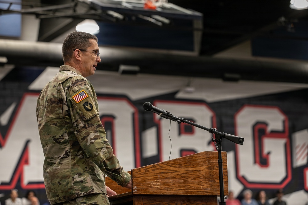 A U.S. Army soldier in OCP (Operational Camouflage Pattern) uniform is speaking at a wooden podium equipped with a microphone. The setting appears to be a school or university gymnasium, identifiable by the large athletic lettering visible on the wall in the background. The soldier wears an American flag patch and an additional unit patch on the left sleeve.