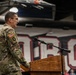 A U.S. Army soldier in OCP (Operational Camouflage Pattern) uniform is speaking at a wooden podium equipped with a microphone. The setting appears to be a school or university gymnasium, identifiable by the large athletic lettering visible on the wall in the background. The soldier wears an American flag patch and an additional unit patch on the left sleeve.
