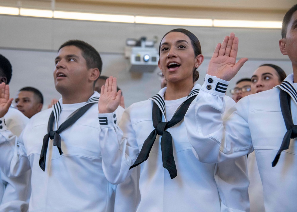 A group of U.S. Navy recruits in white dress uniforms are raising their right hands, appearing to take an oath of enlistment or graduation ceremony. Multiple individuals are visible in formation, all wearing the distinctive white 'Crackerjack' sailor uniforms with black neckerchiefs. The central female recruit appears to be actively reciting the oath with her mouth open.