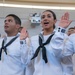 A group of U.S. Navy recruits in white dress uniforms are raising their right hands, appearing to take an oath of enlistment or graduation ceremony. Multiple individuals are visible in formation, all wearing the distinctive white 'Crackerjack' sailor uniforms with black neckerchiefs. The central female recruit appears to be actively reciting the oath with her mouth open.