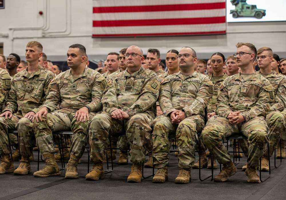 A large group of U.S. Army soldiers in Operational Camouflage Pattern (OCP) uniforms are seated in rows during what appears to be a formal military ceremony or briefing. The soldiers are attentive and facing forward in an indoor venue decorated with a large American flag and a display screen showing a military vehicle. Name tapes are visible on several front-row soldiers including RYAN, FERRO, BONGINO, and CALDWELL.