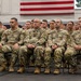 A large group of U.S. Army soldiers in Operational Camouflage Pattern (OCP) uniforms are seated in rows during what appears to be a formal military ceremony or briefing. The soldiers are attentive and facing forward in an indoor venue decorated with a large American flag and a display screen showing a military vehicle. Name tapes are visible on several front-row soldiers including RYAN, FERRO, BONGINO, and CALDWELL.
