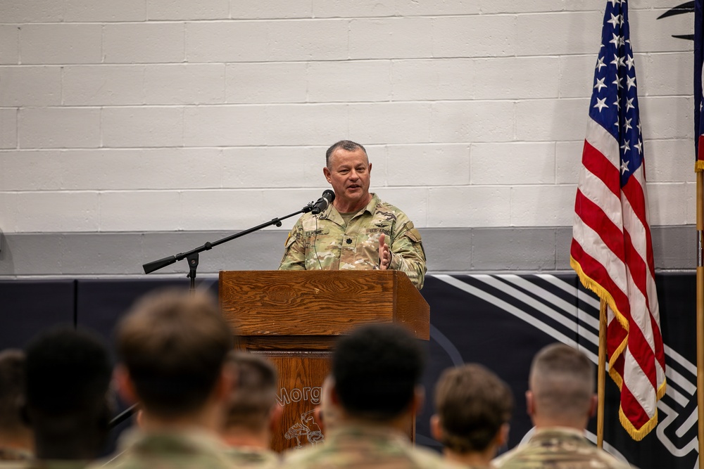 A U.S. Army officer in OCP (Operational Camouflage Pattern) uniform is addressing an audience of uniformed soldiers from a wooden podium. The setting appears to be an indoor military ceremony or briefing, likely in a gymnasium or assembly hall. An American flag and what appears to be a unit guidon or organizational flag are visible to the right of the speaker.