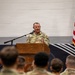A U.S. Army officer in OCP (Operational Camouflage Pattern) uniform is addressing an audience of uniformed soldiers from a wooden podium. The setting appears to be an indoor military ceremony or briefing, likely in a gymnasium or assembly hall. An American flag and what appears to be a unit guidon or organizational flag are visible to the right of the speaker.