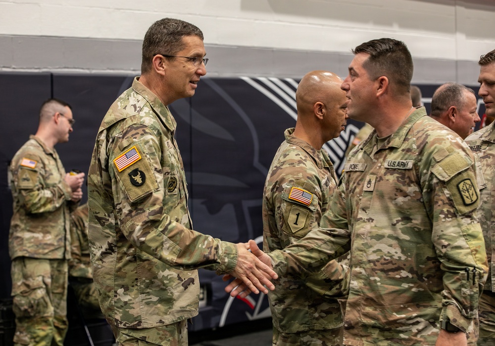 A formal military ceremony is taking place indoors, likely in a gymnasium or drill hall, where a senior officer is shaking hands with soldiers in a receiving line. All personnel are wearing U.S. Army OCP (Operational Camouflage Pattern) uniforms. The setting and handshaking suggest an awards ceremony, promotion, or unit recognition event.