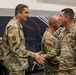 A formal military ceremony is taking place indoors, likely in a gymnasium or drill hall, where a senior officer is shaking hands with soldiers in a receiving line. All personnel are wearing U.S. Army OCP (Operational Camouflage Pattern) uniforms. The setting and handshaking suggest an awards ceremony, promotion, or unit recognition event.