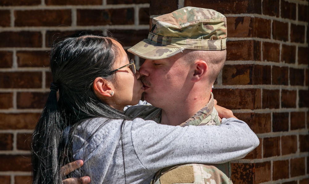 A soldier in military camouflage uniform and patrol cap shares a kiss with a civilian woman against a brick wall background. The moment appears to be an emotional greeting or farewell, with both individuals embracing each other closely. The image is captured in bright daylight with sharp focus on the subjects.