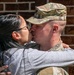 A soldier in military camouflage uniform and patrol cap shares a kiss with a civilian woman against a brick wall background. The moment appears to be an emotional greeting or farewell, with both individuals embracing each other closely. The image is captured in bright daylight with sharp focus on the subjects.