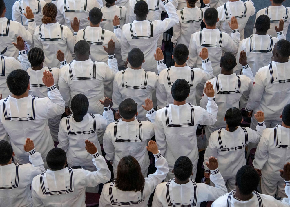 An aerial/overhead view of a large group of individuals in matching white naval uniforms with dark trim and star insignia, raising their right hands in unison. The scene appears to depict a military oath-taking or swearing-in ceremony, likely for U.S. Navy recruits. The uniformity of dress and synchronized gesture strongly suggests a formal induction or graduation ceremony.