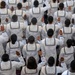 An aerial/overhead view of a large group of individuals in matching white naval uniforms with dark trim and star insignia, raising their right hands in unison. The scene appears to depict a military oath-taking or swearing-in ceremony, likely for U.S. Navy recruits. The uniformity of dress and synchronized gesture strongly suggests a formal induction or graduation ceremony.