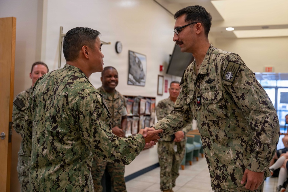 Two U.S. Navy personnel in NWU Type III (green digital camouflage) uniforms are shaking hands in what appears to be an indoor military facility. Several other uniformed personnel are visible in the background, smiling and observing the interaction. The setting suggests a formal or ceremonial occasion such as an award presentation, promotion, or official visit.