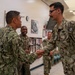 Two U.S. Navy personnel in NWU Type III (green digital camouflage) uniforms are shaking hands in what appears to be an indoor military facility. Several other uniformed personnel are visible in the background, smiling and observing the interaction. The setting suggests a formal or ceremonial occasion such as an award presentation, promotion, or official visit.