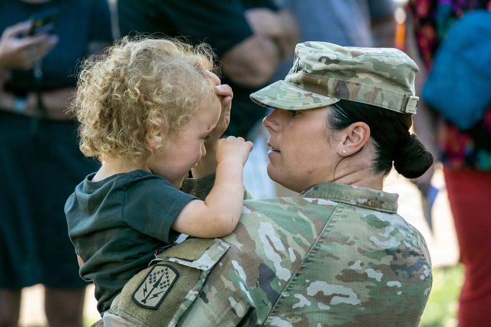 A female soldier in U.S. Army Operational Camouflage Pattern (OCP) uniform holds a young toddler with curly blonde hair. The child appears to be touching or covering the soldier's face in a playful or emotional moment. The scene appears to be an outdoor military homecoming or family event.
