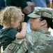 A female soldier in U.S. Army Operational Camouflage Pattern (OCP) uniform holds a young toddler with curly blonde hair. The child appears to be touching or covering the soldier's face in a playful or emotional moment. The scene appears to be an outdoor military homecoming or family event.