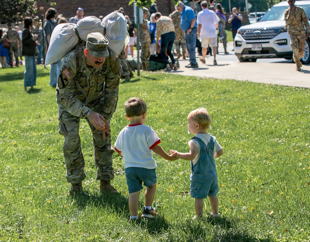 A U.S. Army soldier in MultiCam (OCP) uniform, carrying a large duffel bag over his shoulder, bends down to interact with two young toddlers on a grassy lawn. The scene appears to be a military homecoming or deployment ceremony, with numerous soldiers and civilians gathered in the background. The two small children — one in a white shirt with shorts and one in denim overalls — appear to be greeting the soldier.