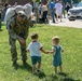 A U.S. Army soldier in MultiCam (OCP) uniform, carrying a large duffel bag over his shoulder, bends down to interact with two young toddlers on a grassy lawn. The scene appears to be a military homecoming or deployment ceremony, with numerous soldiers and civilians gathered in the background. The two small children — one in a white shirt with shorts and one in denim overalls — appear to be greeting the soldier.