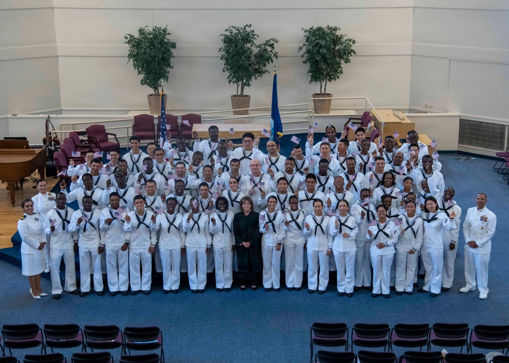 A large group photograph of U.S. Navy personnel in white dress uniforms, gathered in what appears to be an auditorium or ceremonial hall. Most individuals are holding small American flags, suggesting this is a naturalization ceremony or patriotic military event. Two officers in distinct white dress uniforms flank the group on either side, with a civilian in dark clothing positioned at the center front.