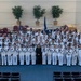 A large group photograph of U.S. Navy personnel in white dress uniforms, gathered in what appears to be an auditorium or ceremonial hall. Most individuals are holding small American flags, suggesting this is a naturalization ceremony or patriotic military event. Two officers in distinct white dress uniforms flank the group on either side, with a civilian in dark clothing positioned at the center front.