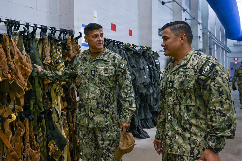 Two U.S. Navy personnel in NWU Type III (woodland digital camouflage) uniforms are conversing inside what appears to be a military equipment storage or gear room. The individual on the left, whose name tape reads 'HICKEY,' is gesturing toward a rack of tactical vests and harnesses. The setting suggests a facility inspection, briefing, or equipment review.
