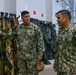 Two U.S. Navy personnel in NWU Type III (woodland digital camouflage) uniforms are conversing inside what appears to be a military equipment storage or gear room. The individual on the left, whose name tape reads 'HICKEY,' is gesturing toward a rack of tactical vests and harnesses. The setting suggests a facility inspection, briefing, or equipment review.