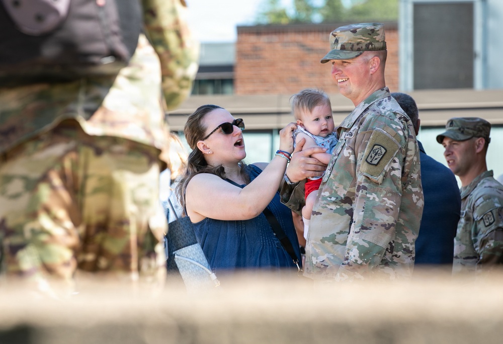 A U.S. Army soldier in OCP (Operational Camouflage Pattern) uniform is holding an infant while a woman, presumably his spouse or partner, interacts joyfully with the baby. The scene appears to be a military homecoming or family day event, with additional soldiers visible in the background. The emotional atmosphere suggests a reunion or celebratory gathering on what appears to be a military installation.