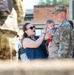 A U.S. Army soldier in OCP (Operational Camouflage Pattern) uniform is holding an infant while a woman, presumably his spouse or partner, interacts joyfully with the baby. The scene appears to be a military homecoming or family day event, with additional soldiers visible in the background. The emotional atmosphere suggests a reunion or celebratory gathering on what appears to be a military installation.