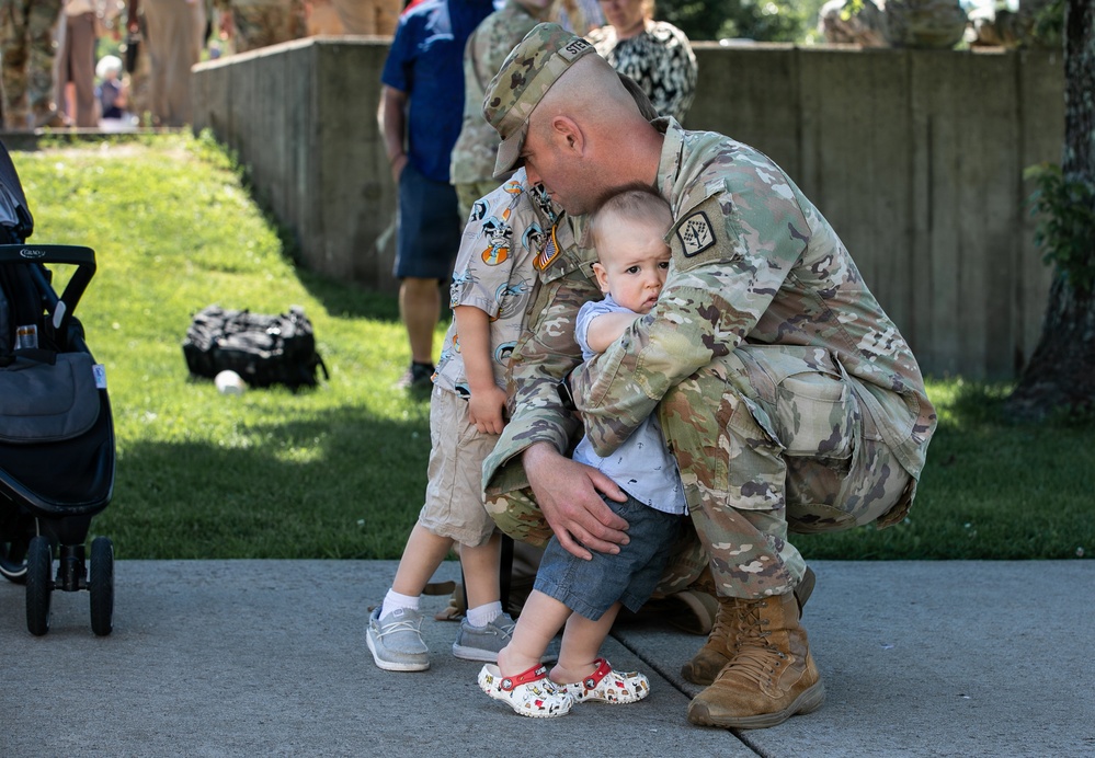 A U.S. Army soldier in OCP (Operational Camouflage Pattern) uniform is crouched down embracing two young children, likely his own, in what appears to be a homecoming or farewell event. The soldier is wearing a patrol cap and tan combat boots, holding a baby while an older toddler stands beside him. The outdoor setting with other military personnel and families visible in the background suggests a unit deployment or redeployment ceremony.