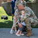 A U.S. Army soldier in OCP (Operational Camouflage Pattern) uniform is crouched down embracing two young children, likely his own, in what appears to be a homecoming or farewell event. The soldier is wearing a patrol cap and tan combat boots, holding a baby while an older toddler stands beside him. The outdoor setting with other military personnel and families visible in the background suggests a unit deployment or redeployment ceremony.