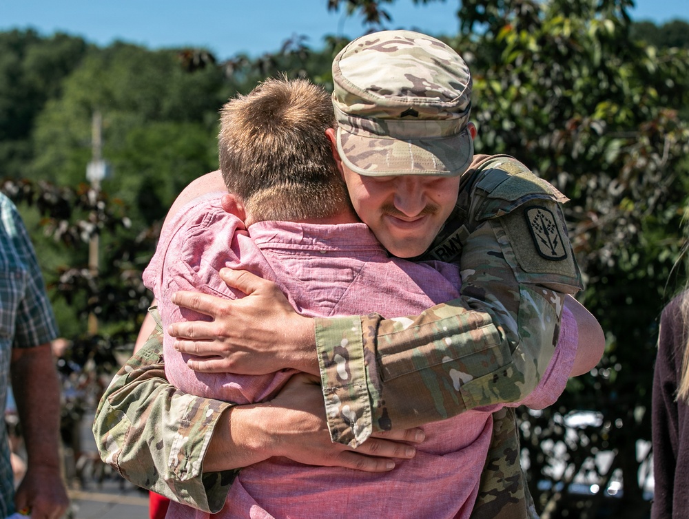 A U.S. Army soldier in OCP (Operational Camouflage Pattern) uniform is embracing a civilian wearing a pink shirt in what appears to be a homecoming or farewell event. The soldier is smiling warmly during the hug, suggesting an emotional reunion or departure moment. The outdoor setting with trees and blue sky in the background indicates a daytime public gathering.