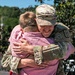 A U.S. Army soldier in OCP (Operational Camouflage Pattern) uniform is embracing a civilian wearing a pink shirt in what appears to be a homecoming or farewell event. The soldier is smiling warmly during the hug, suggesting an emotional reunion or departure moment. The outdoor setting with trees and blue sky in the background indicates a daytime public gathering.