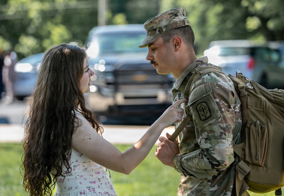 A young woman in a floral dress interacts closely with a male soldier in U.S. Army OCP (Operational Camouflage Pattern) uniform who is wearing a backpack. The woman appears to be adjusting or touching the soldier's uniform or gear, suggesting a farewell or reunion moment. The setting is an outdoor area with vehicles and trees visible in the blurred background.