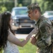 A young woman in a floral dress interacts closely with a male soldier in U.S. Army OCP (Operational Camouflage Pattern) uniform who is wearing a backpack. The woman appears to be adjusting or touching the soldier's uniform or gear, suggesting a farewell or reunion moment. The setting is an outdoor area with vehicles and trees visible in the blurred background.