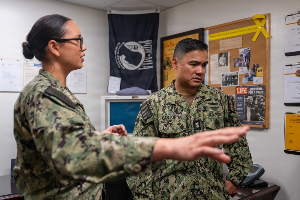 Two U.S. Navy personnel in digital camouflage uniforms (NWU Type III) are engaged in a conversation inside what appears to be a military office. The male sailor's name tape reads 'CANTORNA' and his uniform displays U.S. Navy insignia. The room contains a POW/MIA flag, a bulletin board with photographs and documents, and a vintage LIFE magazine cover.