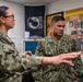 Two U.S. Navy personnel in digital camouflage uniforms (NWU Type III) are engaged in a conversation inside what appears to be a military office. The male sailor's name tape reads 'CANTORNA' and his uniform displays U.S. Navy insignia. The room contains a POW/MIA flag, a bulletin board with photographs and documents, and a vintage LIFE magazine cover.