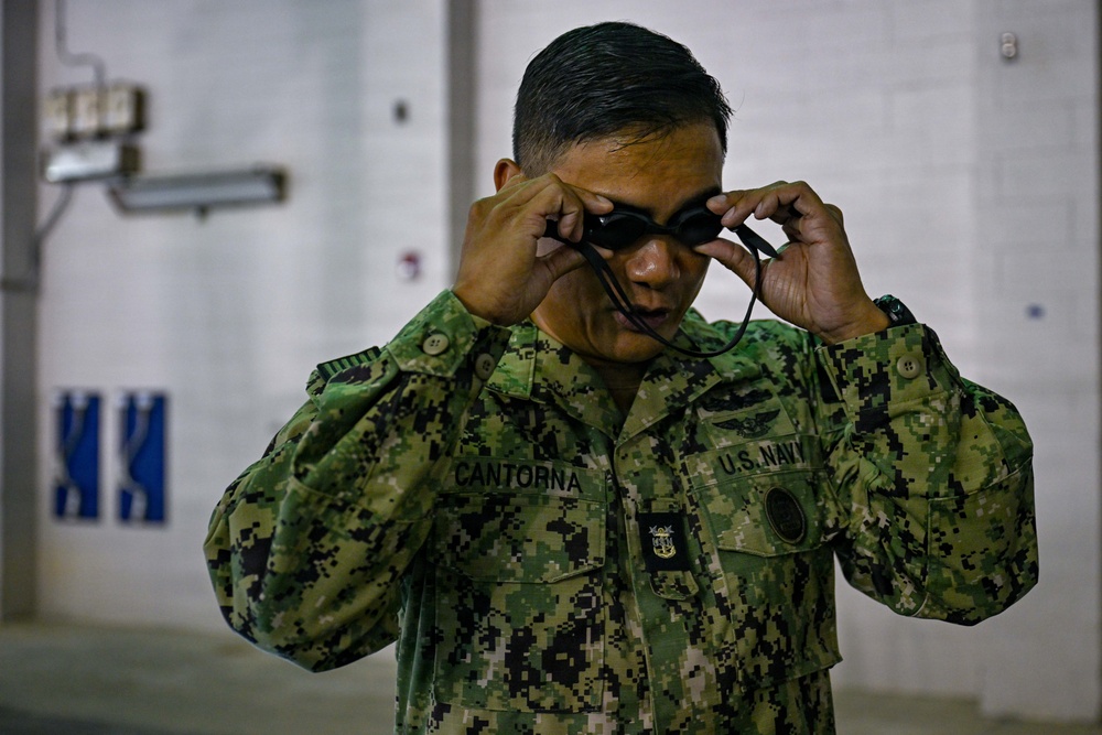 A U.S. Navy service member named Cantorna is adjusting swim goggles over their eyes. The individual is wearing the Navy Working Uniform (NWU) Type III in green digital camouflage pattern. The setting appears to be an indoor facility, likely near a swimming pool or aquatic training area.