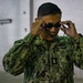 A U.S. Navy service member named Cantorna is adjusting swim goggles over their eyes. The individual is wearing the Navy Working Uniform (NWU) Type III in green digital camouflage pattern. The setting appears to be an indoor facility, likely near a swimming pool or aquatic training area.