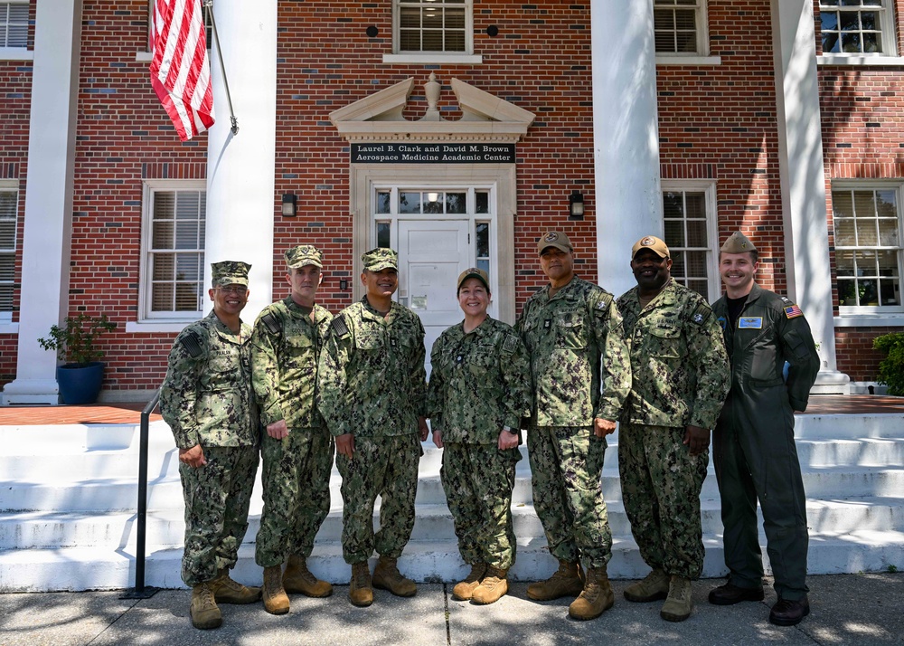 Seven military personnel pose for a group photo in front of the Laurel B. Clark and David M. Brown Aerospace Medicine Academic Center, a red brick building with white colonial columns. Six individuals are wearing U.S. Navy Working Uniform (NWU Type III) camouflage, while one person on the far right wears an olive green flight suit. An American flag is visible on a flagpole to the upper left of the building.