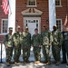 Seven military personnel pose for a group photo in front of the Laurel B. Clark and David M. Brown Aerospace Medicine Academic Center, a red brick building with white colonial columns. Six individuals are wearing U.S. Navy Working Uniform (NWU Type III) camouflage, while one person on the far right wears an olive green flight suit. An American flag is visible on a flagpole to the upper left of the building.