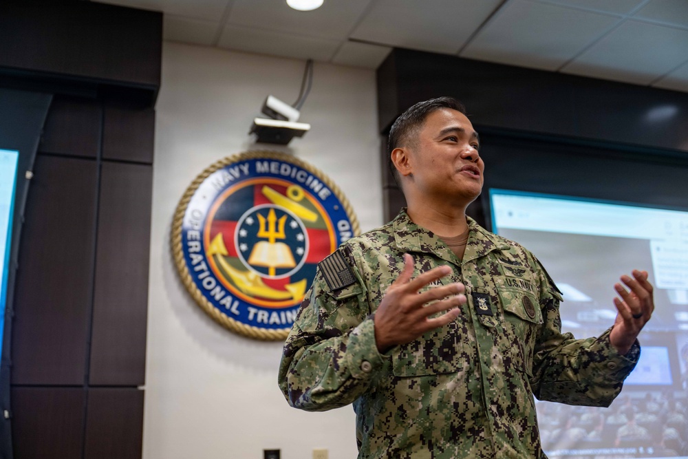 A U.S. Navy service member in digital camouflage (NWU Type III) uniform is presenting or speaking in what appears to be a briefing room. Behind them is the official seal of Navy Medicine Operational Training Command (NMOTC) mounted on the wall. A projection screen is visible to the right, suggesting an active presentation or training session.