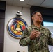A U.S. Navy service member in digital camouflage (NWU Type III) uniform is presenting or speaking in what appears to be a briefing room. Behind them is the official seal of Navy Medicine Operational Training Command (NMOTC) mounted on the wall. A projection screen is visible to the right, suggesting an active presentation or training session.