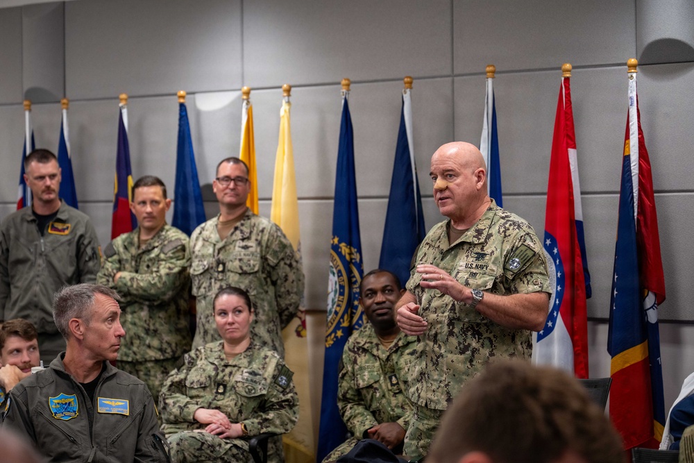 A senior U.S. Navy officer in camouflage uniform is addressing a group of military personnel in what appears to be a formal briefing or meeting room. Multiple service members from various branches are present, seated and standing, with a row of U.S. state and military flags displayed in the background. The setting suggests an official military gathering or command briefing.