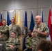 A senior U.S. Navy officer in camouflage uniform is addressing a group of military personnel in what appears to be a formal briefing or meeting room. Multiple service members from various branches are present, seated and standing, with a row of U.S. state and military flags displayed in the background. The setting suggests an official military gathering or command briefing.