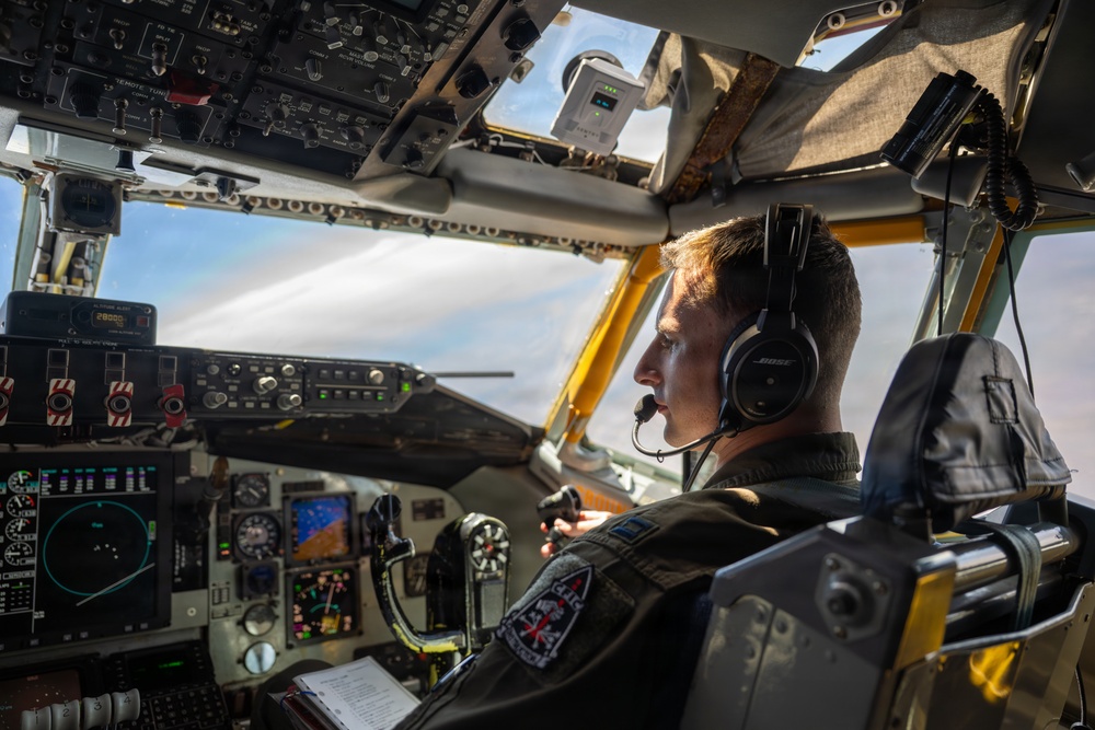 A military pilot is seated in the cockpit of a large aircraft, likely a tanker or transport plane, during flight. The pilot is wearing a dark flight suit, a Bose aviation headset with boom microphone, and has a squadron patch visible on his shoulder. The cockpit features an extensive array of analog and digital instrumentation panels.